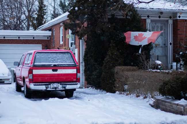 A red truck parked in a snowy Toronto front yard with a Canadian flag waving by the brick house.